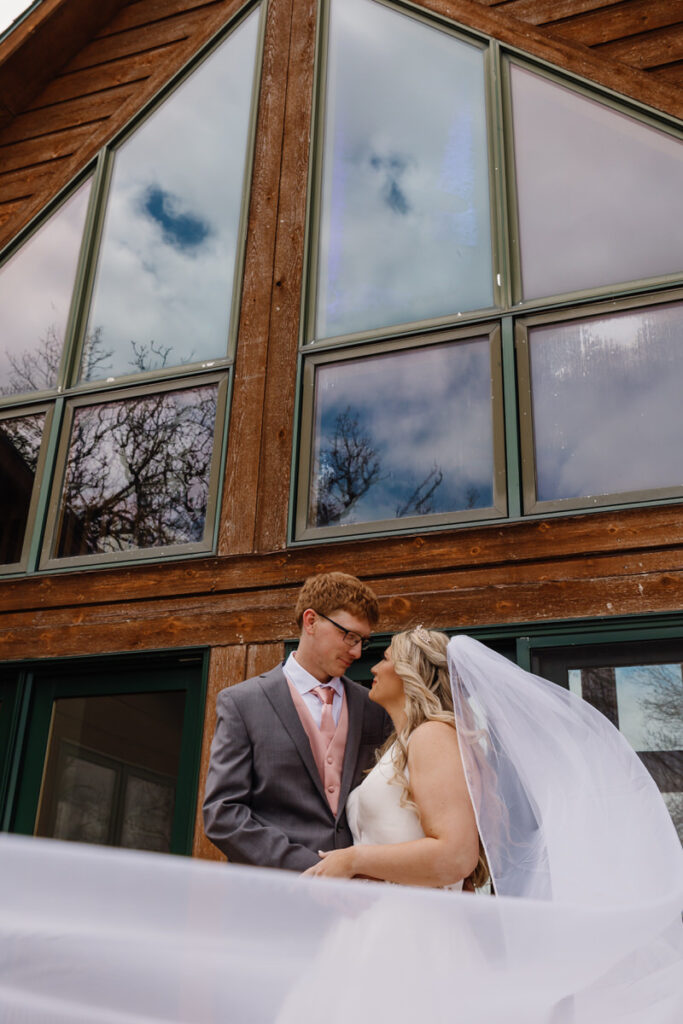 A newlywed couple standing outside an A-frame building with a veil flowing in front of them 