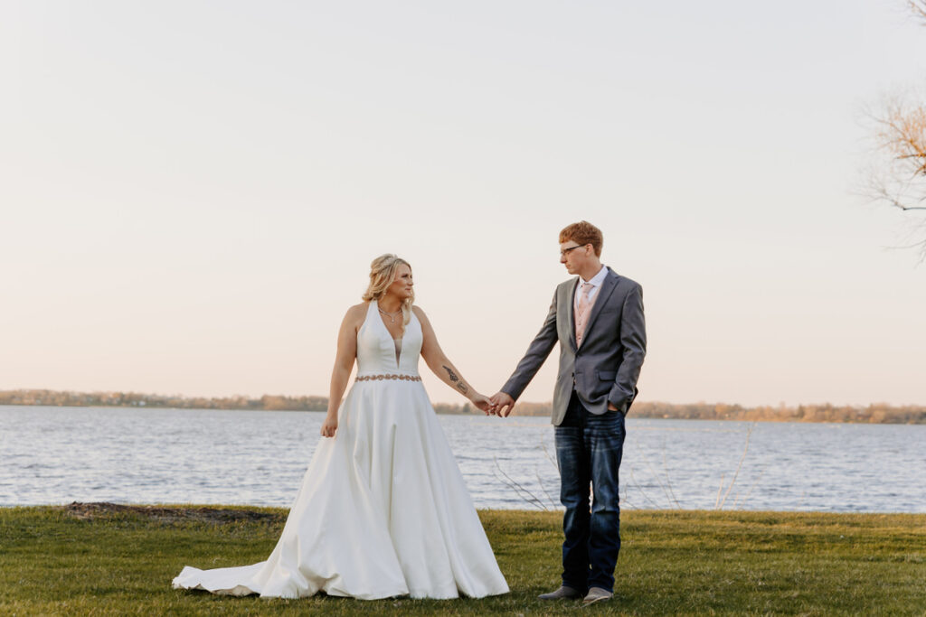 A newlywed couple holding hands and looking at each other in front of a lake 