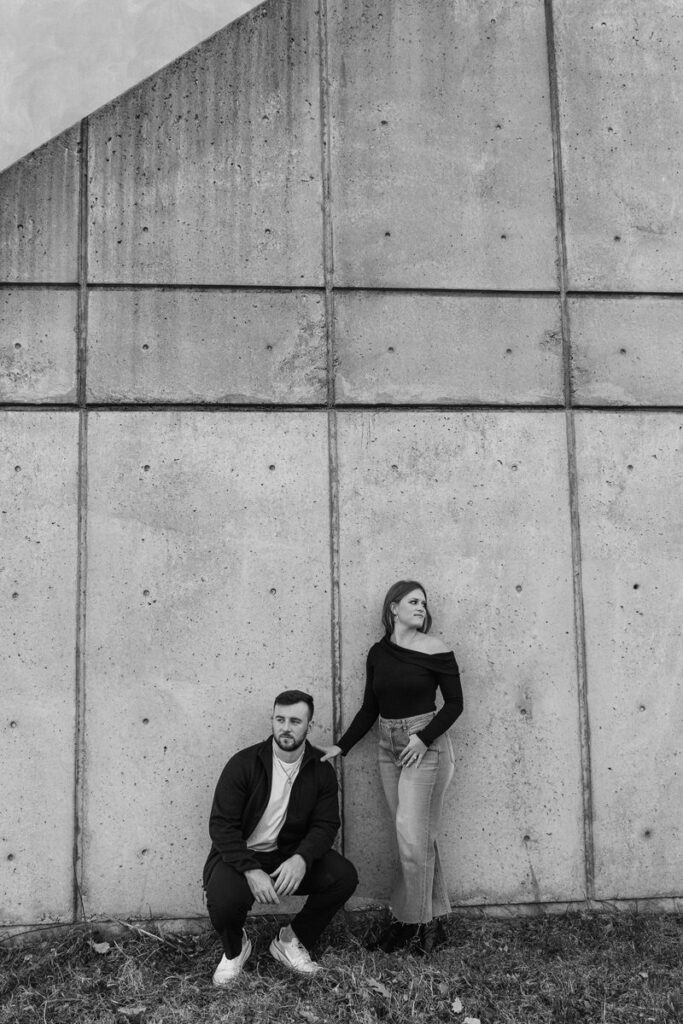 A couple standing against a concrete wall with one of them crouching down 