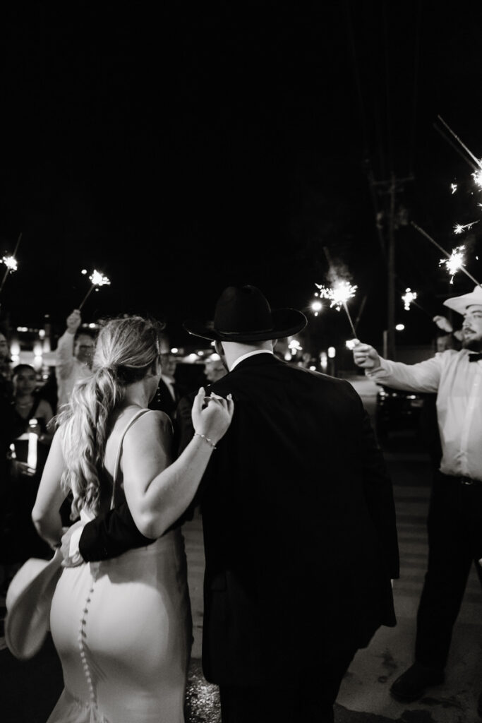 A newlywed couple walking through a tunnel of their guests holding up sparklers 
