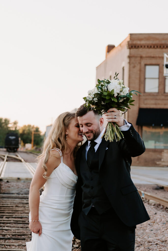 A newlywed couple standing close and laughing as one of them holds up a bouquet of flowers 